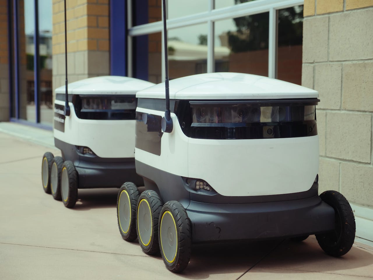 who-we-are Two autonomous delivery robots positioned outside a modern building, showcasing innovation in robotics and mobility.